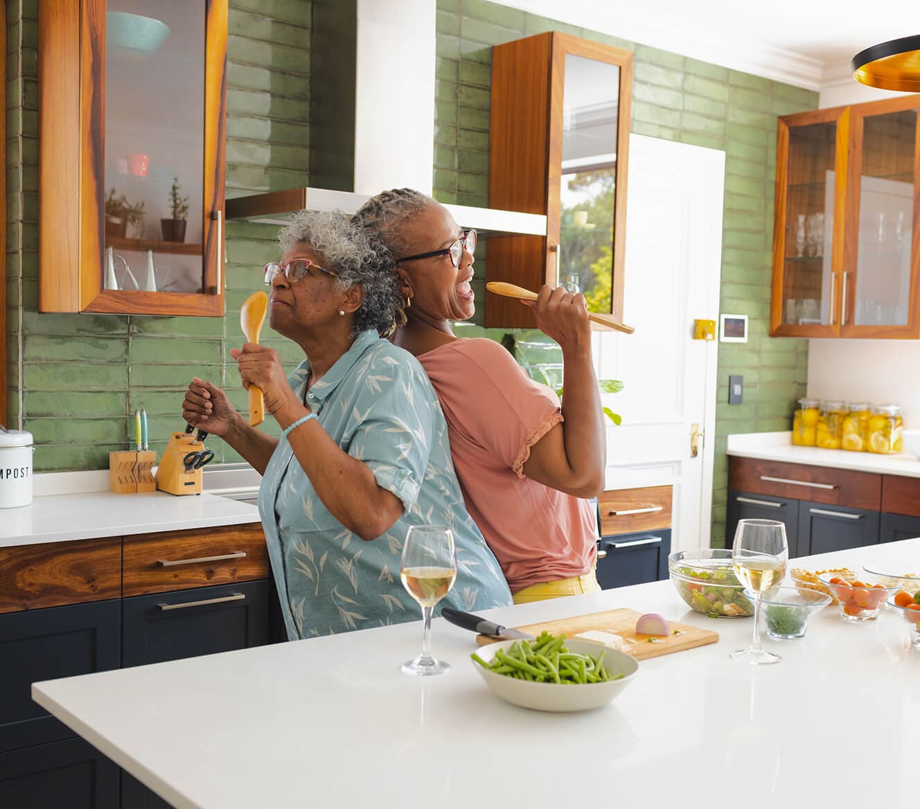 Two joyful older women in a modern kitchen, standing back-to-back while singing into wooden cooking utensils like microphones. They are surrounded by fresh ingredients and glasses of white wine, enjoying a fun and lighthearted moment while cooking togethe Two joyful older women in a modern kitchen, standing back-to-back while singing into wooden cooking utensils like microphones. They are surrounded by fresh ingredients and glasses of white wine enjoying a fun and lighthearted moment while cooking together
