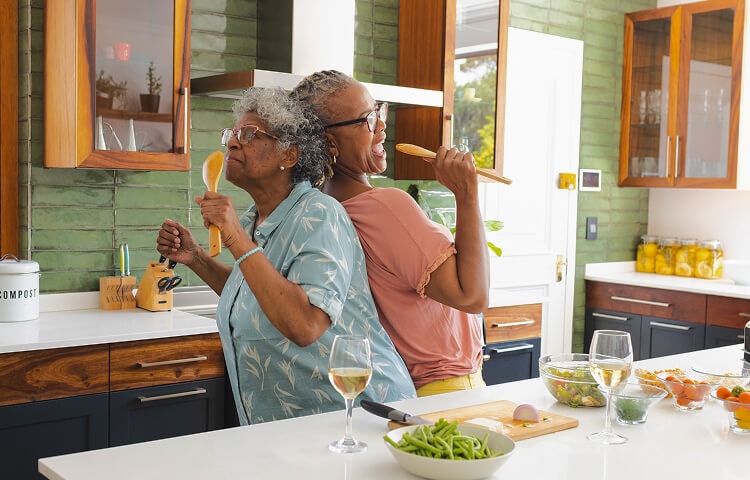 Two joyful older women in a modern kitchen, standing back-to-back while singing into wooden cooking utensils like microphones. They are surrounded by fresh ingredients and glasses of white wine, enjoying a fun and lighthearted moment while cooking togethe Two joyful older women in a modern kitchen, standing back-to-back while singing into wooden cooking utensils like microphones. They are surrounded by fresh ingredients and glasses of white wine enjoying a fun and lighthearted moment while cooking together