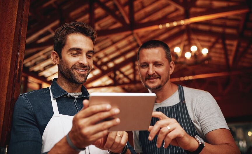 A flood sensor is mounted under a sink with visible plumbing, detecting leaks to help prevent damage. Two men in aprons smile while reviewing lighting information on a tablet, representing small business owners using smart tools.