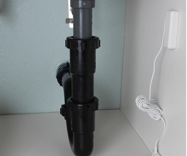 A flood sensor is mounted under a sink with visible plumbing, detecting leaks to help prevent damage. A flood sensor is mounted under a sink with visible plumbing, detecting leaks to help prevent damage.