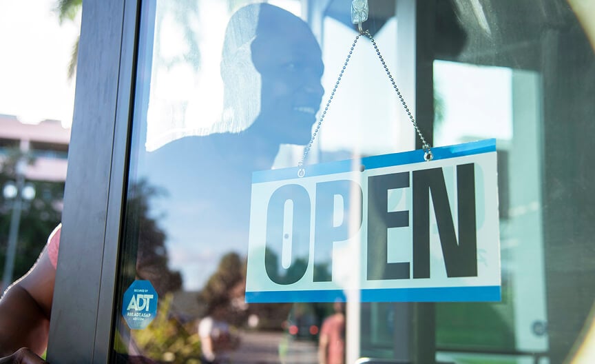 ADT Command Panel A hanging “Open” sign on a glass door with an ADT security sticker, and the reflection of a smiling customer.