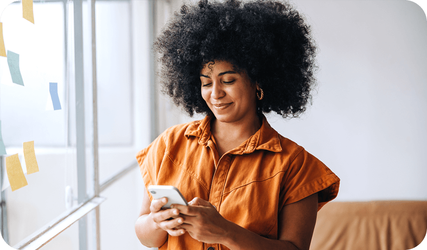 Woman smiling while using smartphone Smiling woman with curly hair in an orange shirt using her smartphone near a window.