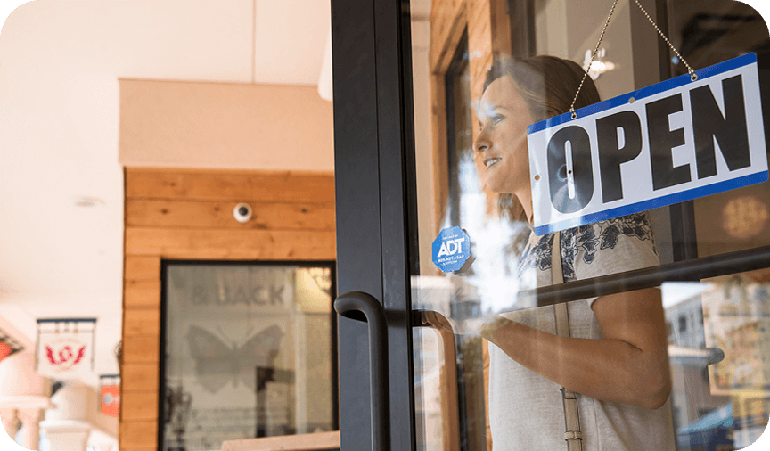 “Open” sign hanging on a glass storefront door with an ADT security sticker and a woman visible inside. Open sign on storefront door
