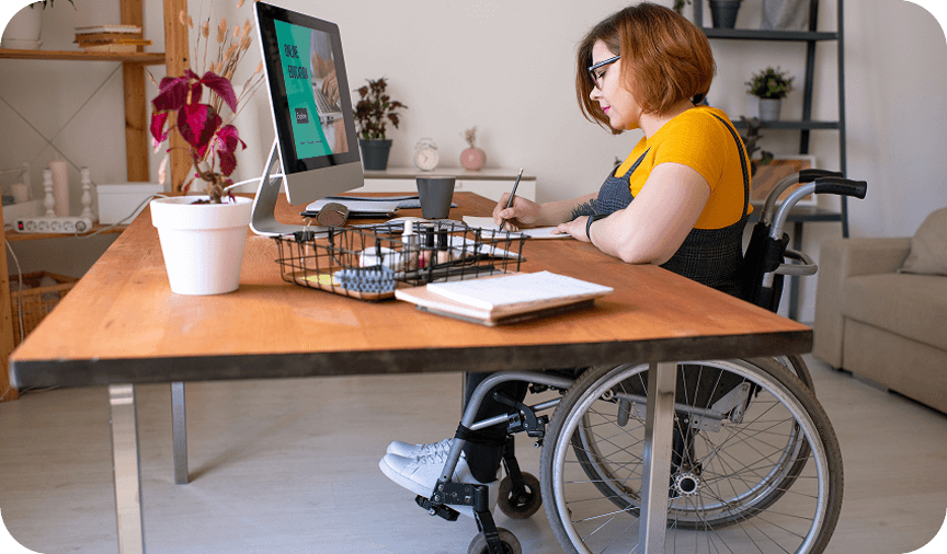 Woman working at desk in wheelchair Woman using a wheelchair working at a desk, writing in a notebook beside a desktop computer.