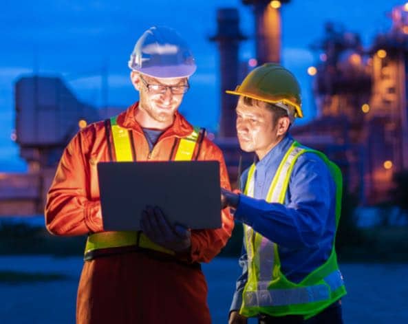 Refinery workers looking at laptop outdoors in twilight